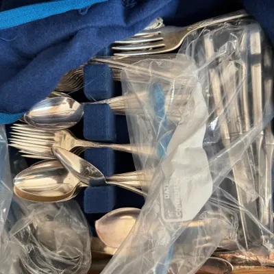 Assorted silver-plated flatware set stored in a blue felt-lined drawer insert, with some pieces wrapped in protective plastic bags.