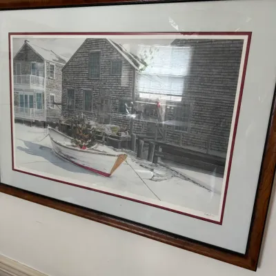 Framed and matted art print depicting a winter harbor scene with a dory boat and shingled coastal buildings, housed in a wood-grain frame.