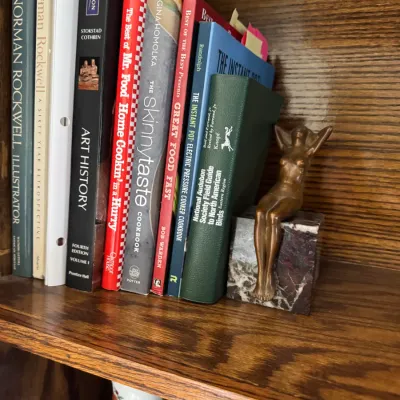 Sale Photo Thumbnail #166: Art Deco style bronze bookend featuring a seated female figure on a marble base, supporting a row of books on a wooden shelf.