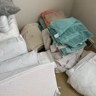 Assorted household linens including stacks of bath towels in teal, pink, and beige, alongside various bed pillows and white bedding.