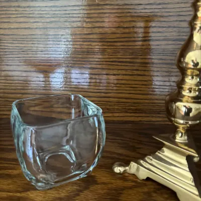 Sale Photo Thumbnail #163: Small square clear glass votive holder shown next to a polished brass candlestick base on a wooden surface.