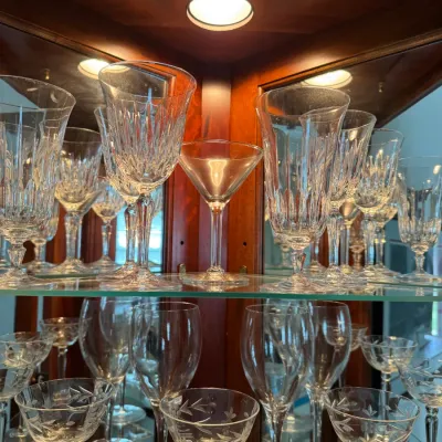 Collection of vintage crystal stemware, including cut-glass goblets and etched coupe glasses, displayed on glass shelves inside a lighted...
