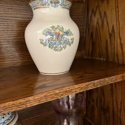Sale Photo Thumbnail #171: Lenox porcelain vase featuring a floral and fruit basket motif with gold trim, displayed on a wooden shelf.