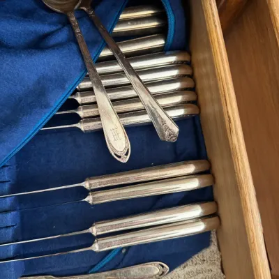Vintage silver-plated flatware set, including knives and spoons, stored in a blue tarnish-resistant cloth roll inside a wooden drawer.
