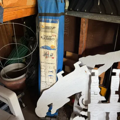 First Up straight wall gazebo in its blue carrying case, stored alongside white wooden yard decorations and gardening supplies.