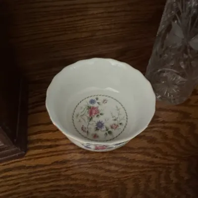 Sale Photo Thumbnail #191: Small porcelain bowl with a scalloped rim, featuring a delicate floral pattern in pink and purple on the interior bottom, displayed on a wooden shelf next to a cut crystal vase.