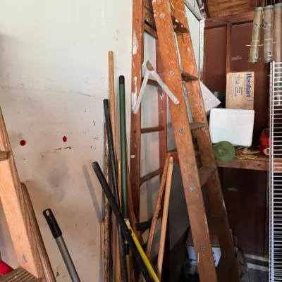 Vintage wooden A-frame stepladder with paint splatters, shown alongside an assortment of long-handled garden tools and brooms in a garage.