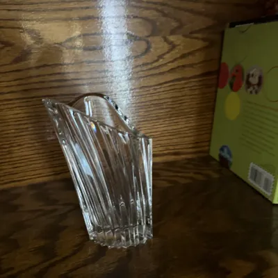 Sale Photo Thumbnail #144: Small clear cut-glass vase with a ribbed texture and asymmetrical, sloped rim, displayed on a wooden shelf.