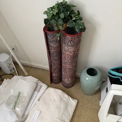 Assorted home decor items including two rolled patterned runner rugs, a light blue ceramic vase, a faux potted plant, and folded white...