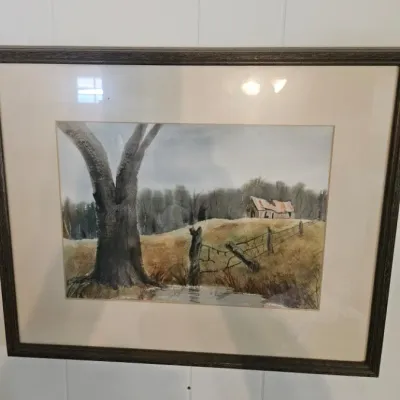 Framed watercolor painting depicting a rustic rural landscape with a large tree, fence line, and a distant barn, presented in a dark wood...