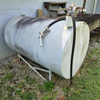 Large horizontal fuel storage tank with a manual hand pump, hose, and nozzle, showing weathered wear from outdoor use.