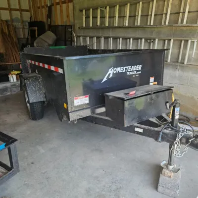 Black Homesteader utility trailer with a front-mounted toolbox, showing some wear and mud splatters from use.
