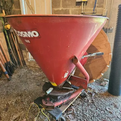 Cosmo 500 pull-behind fertilizer spreader with a red cone-shaped hopper, showing some wear and rust from use.