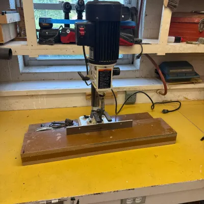 Benchtop mortising machine mounted on a wooden base, showing some signs of use, situated on a yellow workshop table.