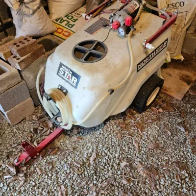 NorthStar tow-behind trailer sprayer featuring a large white poly tank, integrated pump, and pressure gauge, showing some wear from...
