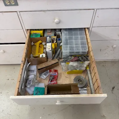 An open workbench drawer containing various hardware supplies, including nails, screws in a plastic organizer, springs, and small tools.