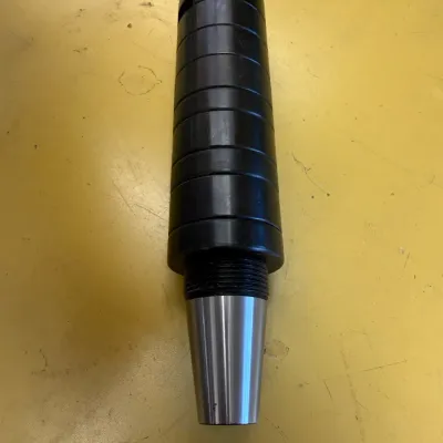 Industrial metal tool holder or arbor with a tapered shank and a stack of black spacer rings, shown on a yellow surface.