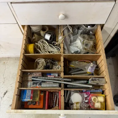 An open wooden workbench drawer with adjustable dividers, filled with various hardware items including springs, rope, tape, and packaged...