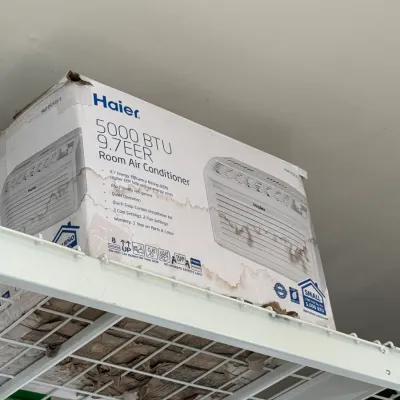 Sale Photo Thumbnail #212: Haier 5000 BTU room air conditioner in its original box, showing some water damage to the packaging, stored on a wire shelf.