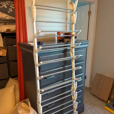 Sale Photo Thumbnail #150: Over-the-door multi-tier shoe rack with white plastic brackets and grey metal rods, leaning against a dark grey wooden dresser.