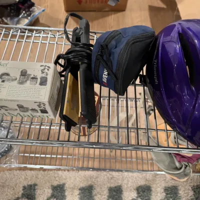Sale Photo Thumbnail #118: Assorted household items on a wire shelf, including a purple Triumph bicycle helmet, an Avenir zippered pouch, a black hair straightener, and a boxed PotMaker kit.