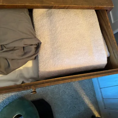 Sale Photo Thumbnail #146: Assorted linens, including a folded white towel and grey sheets, stored inside a vintage wooden dresser drawer.