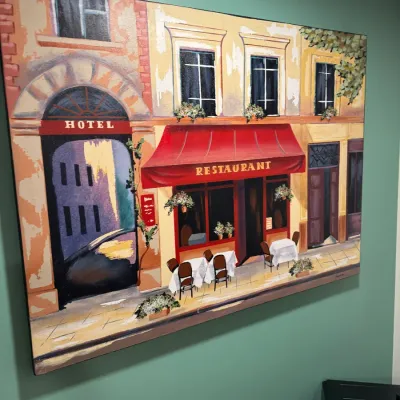 Large unframed canvas painting depicting a European street scene with a hotel and a restaurant with outdoor seating.