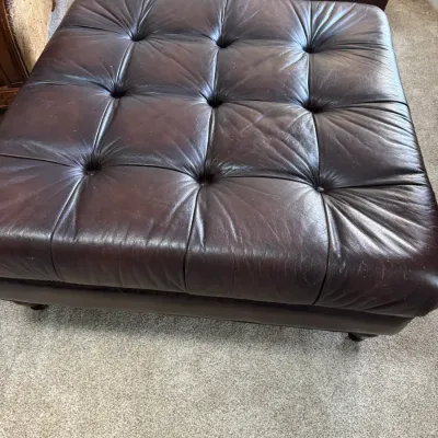 Large square tufted leather ottoman in dark brown, showing some light wear and creasing consistent with use.