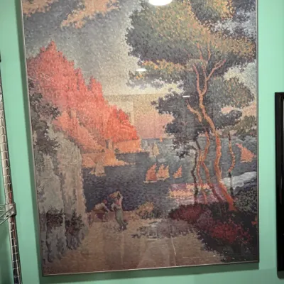 Large framed pointillist-style print depicting a coastal landscape with sailboats, figures, and trees, housed in a simple metal frame.