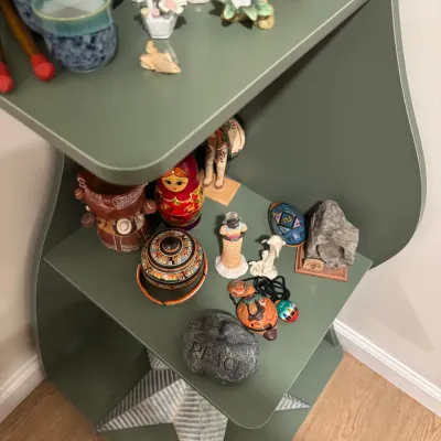 Sale Photo Thumbnail #281: Assorted small collectibles and decorative items, including a nesting doll, pottery, and figurines, displayed on a green multi-tiered corner shelf.