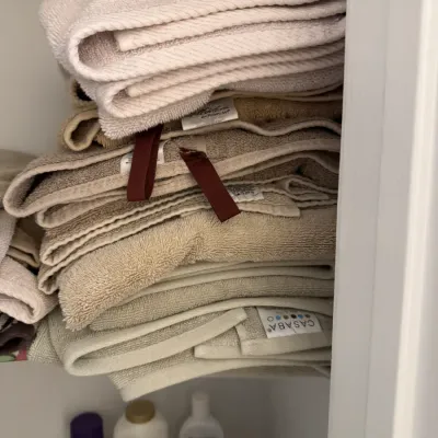 Sale Photo Thumbnail #154: Stack of assorted bath towels in neutral tones, including brands like Casaba, stored on a closet shelf.