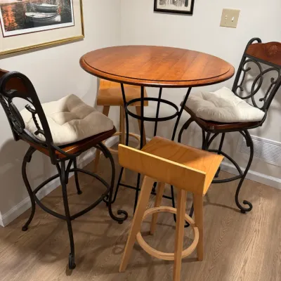 Round wooden pub table with a black metal base, paired with two matching wrought iron barstools with white cushions and two light wood...