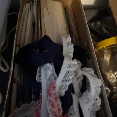 Sale Photo Thumbnail #194: Assorted vintage sewing notions, including white eyelet lace trim, pink decorative trim, and dark blue fabric, stored inside a cardboard box.