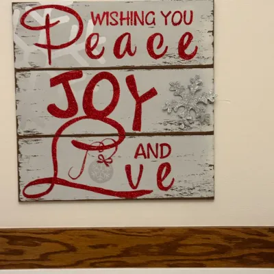 Rustic wooden Christmas wall sign featuring the message "Wishing You Peace Joy and Love" in red glitter lettering, accented...