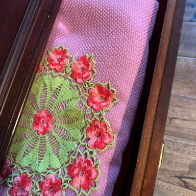 Vintage crocheted doily with green and red floral details, displayed on a purple woven textile inside a wooden drawer.