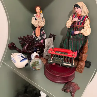 Sale Photo Thumbnail #131: Assorted vintage travel souvenirs and figurines, including European dolls in traditional dress, a San Francisco cable car music box, a Delft-style ceramic shoe, and a carved dragon.
