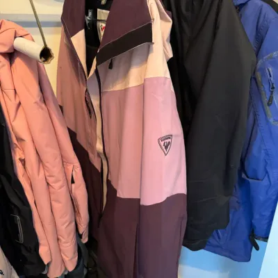 Rossignol women's color-blocked ski jacket in shades of purple, pink, and cream, hanging alongside other winter outerwear.