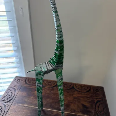 Handcrafted folk art giraffe sculpture constructed from recycled green aluminum soda cans and wire, standing on a carved wooden table.