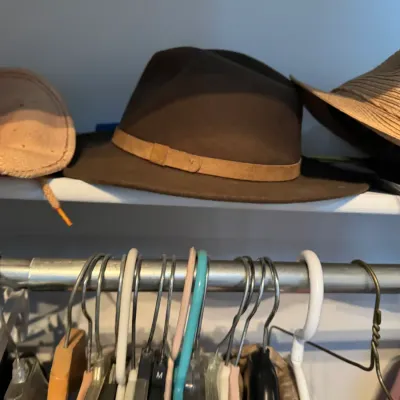 Sale Photo Thumbnail #173: Brown felt fedora hat with a tan leather band, displayed on a closet shelf above a clothing rack.