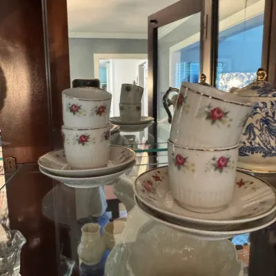 Vintage porcelain teacups and saucers featuring a delicate pink rose pattern and gold trim, displayed inside a glass-front china cabinet.