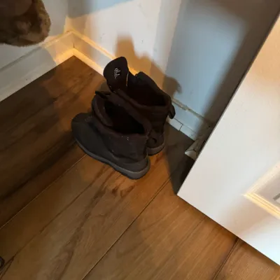 Sale Photo Thumbnail #174: Pair of dark brown or black winter boots sitting on a wood-look floor in a corner.