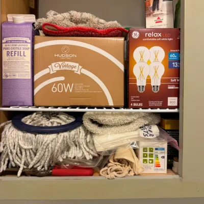 Sale Photo Thumbnail #233: Assorted household supplies including Dr. Bronner's lavender soap, Hudson Lighting vintage 60W bulbs, GE Relax LED bulbs, and a mop head stored on wire shelving.