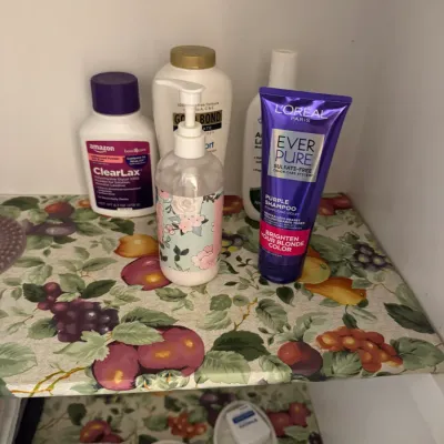 Sale Photo Thumbnail #152: Assorted personal care and hygiene products, including L'Oreal Ever Pure purple shampoo, Gold Bond lotion, and ClearLax, arranged on a fruit-patterned shelf liner.