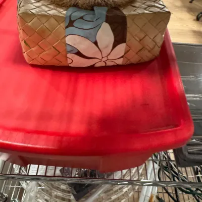 Woven pandanus leaf gift box decorated with a floral fabric band and a coconut fiber rosette, resting on a red plastic storage bin.