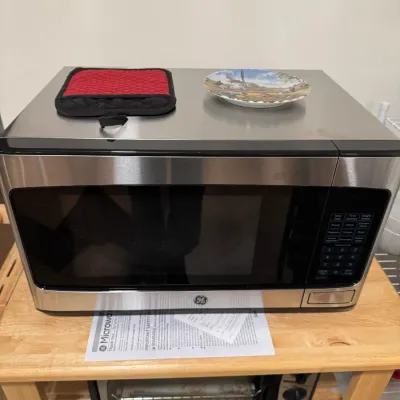 Sale Photo Thumbnail #135: GE stainless steel countertop microwave oven, shown sitting on a wooden cart with its user manual underneath.