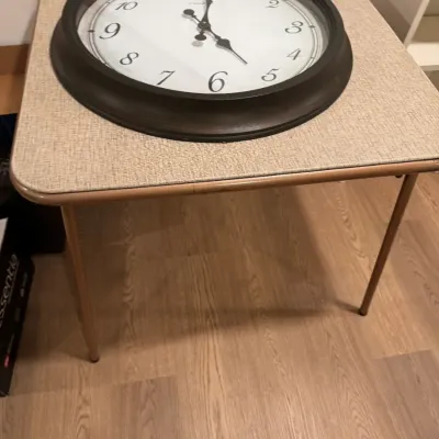 Sale Photo Thumbnail #276: Large round wall clock with a dark brown frame and white face, displayed on a vintage square folding card table with a textured beige top.