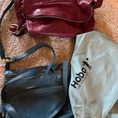 Two Hobo the Original leather handbags, including a burgundy foldover crossbody bag and a blue multi-zipper shoulder bag, shown with a...