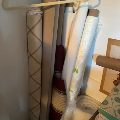 Sale Photo Thumbnail #198: Assorted rolls of fabric and craft paper, including a diamond-patterned beige textile and a striped maroon material, stored upright in a closet.