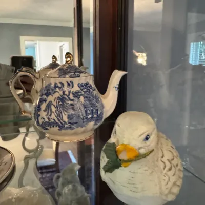Vintage Blue Willow pattern ceramic teapot with gold trim, displayed alongside a small white bird figurine inside a glass curio cabinet.
