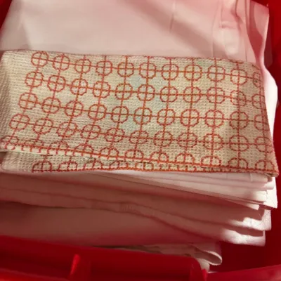 Stack of vintage pink linens and a hand-embroidered kitchen towel with a geometric orange pattern, stored in a red plastic bin.
